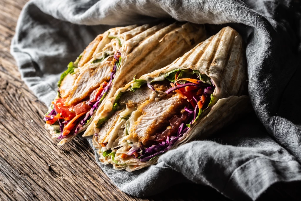 Połówki wrapa z grillowanym kurczakiem, świeżymi warzywami takimi jak sałata, kapusta i papryka, owinięte w cienki placek, ułożone na lnianej ściereczce na drewnianym stole.