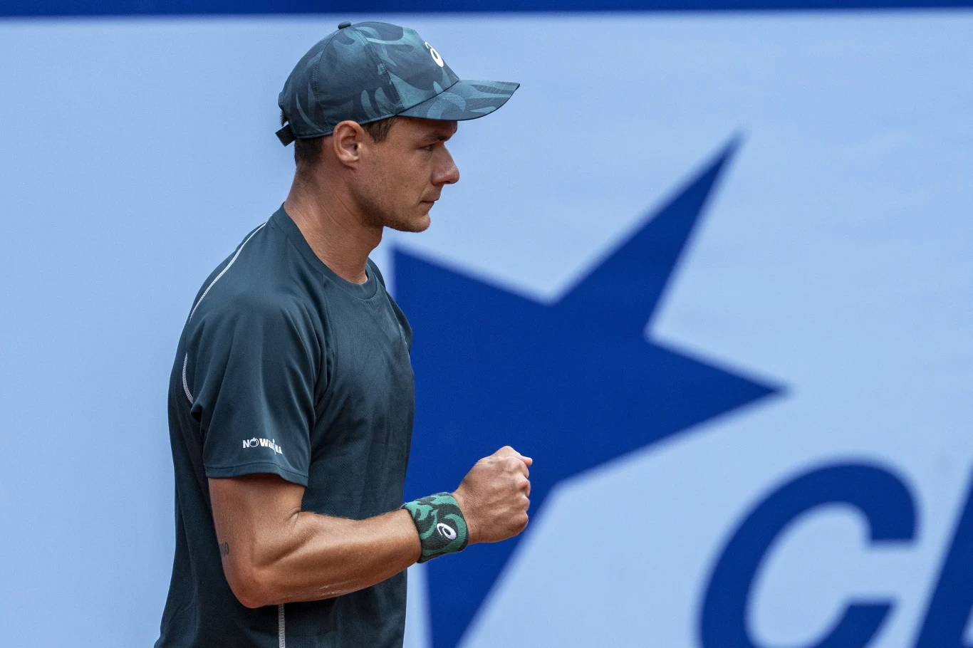 Sportowiec ubrany w ciemny strój i czapkę w geście triumfu lub determinacji, zaciśnięta pięść, w tle fragment logo z dużą niebieską gwiazdą.