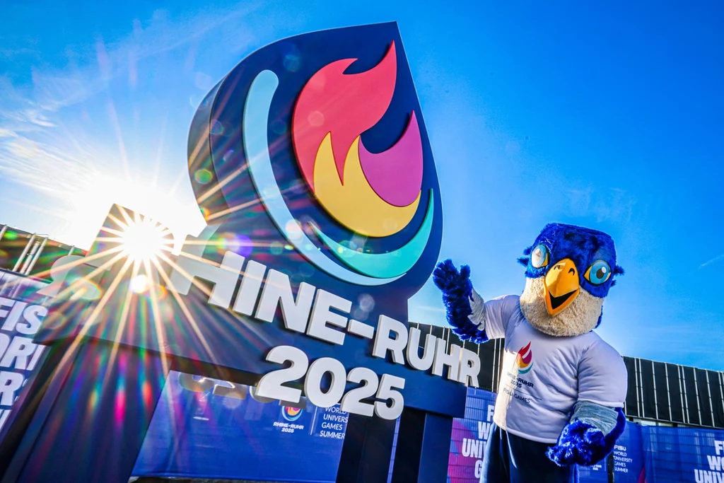 Logotyp tegorocznej uniwersjady w Niemczech Maskotka w stroju niebieskiego ptaka pozuje obok dużego znaku z logo i napisem „RHINE-RUHR 2025” na tle nieba i promieni słońca. W tle widoczna infrastruktura wydarzenia oraz wyraźne barwy sportowe.