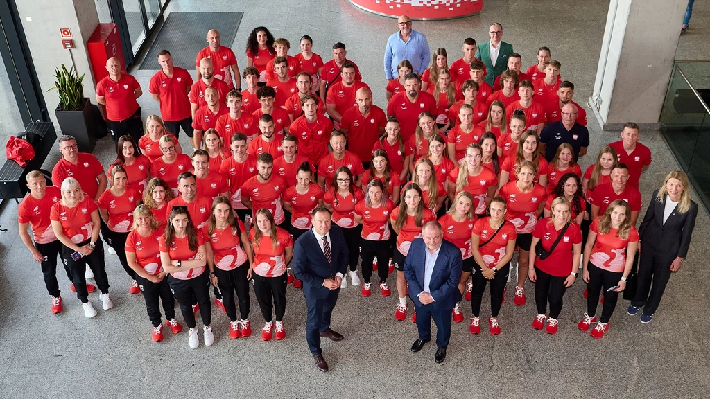 Polska reprezentacja na Uniwersjadę w Niemczech Grupa osób ubranych w jednakowe czerwone koszulki sportowe, ustawionych na przestronnym korytarzu, większość patrzy w stronę aparatu, na przedzie kilku mężczyzn w garniturach.
