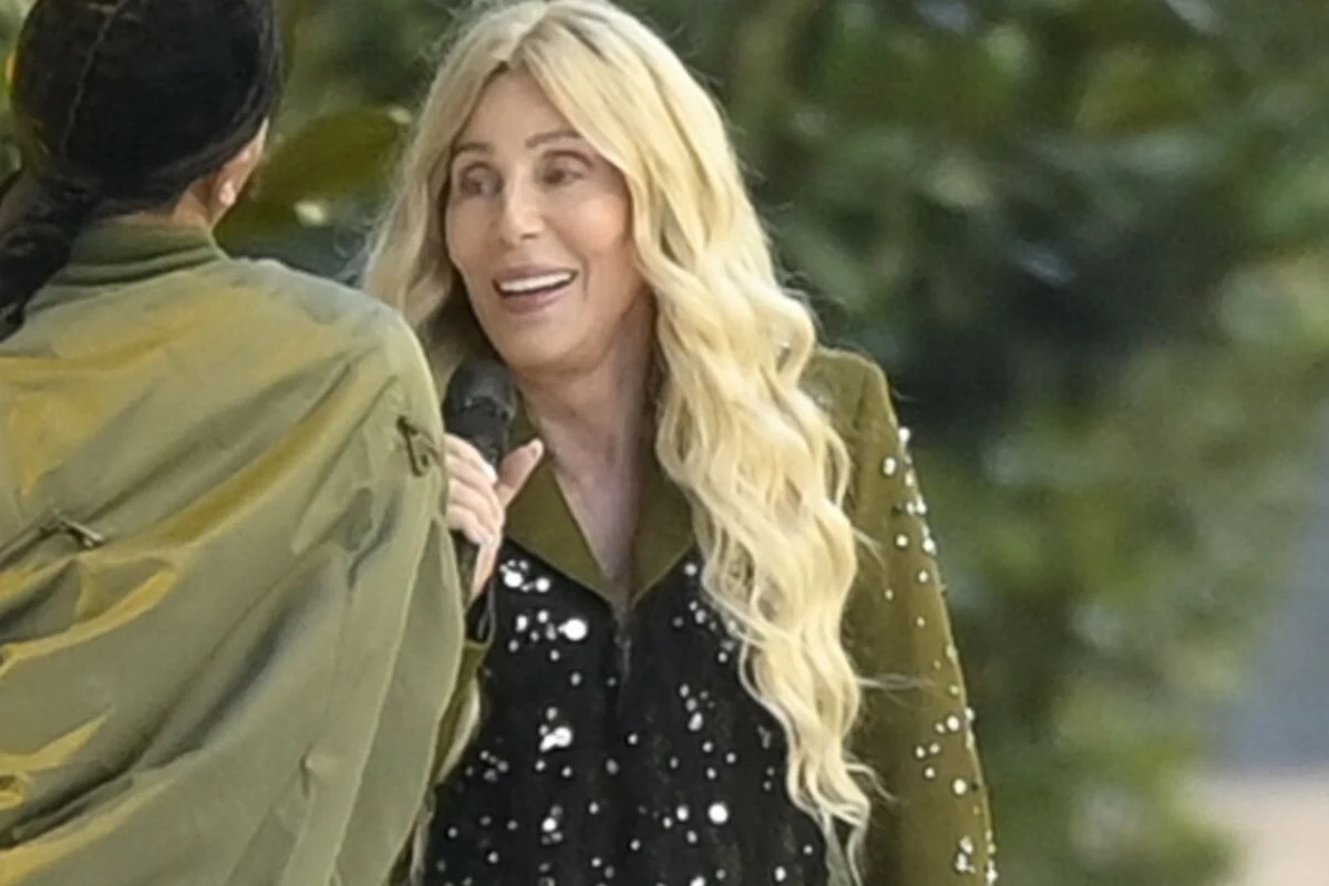 Cher wciąż zachwyca w wieku 79 lat Kobieta o długich blond włosach i jasnym uśmiechu trzyma mikrofon i rozmawia z inną osobą, w tle zielone liście.