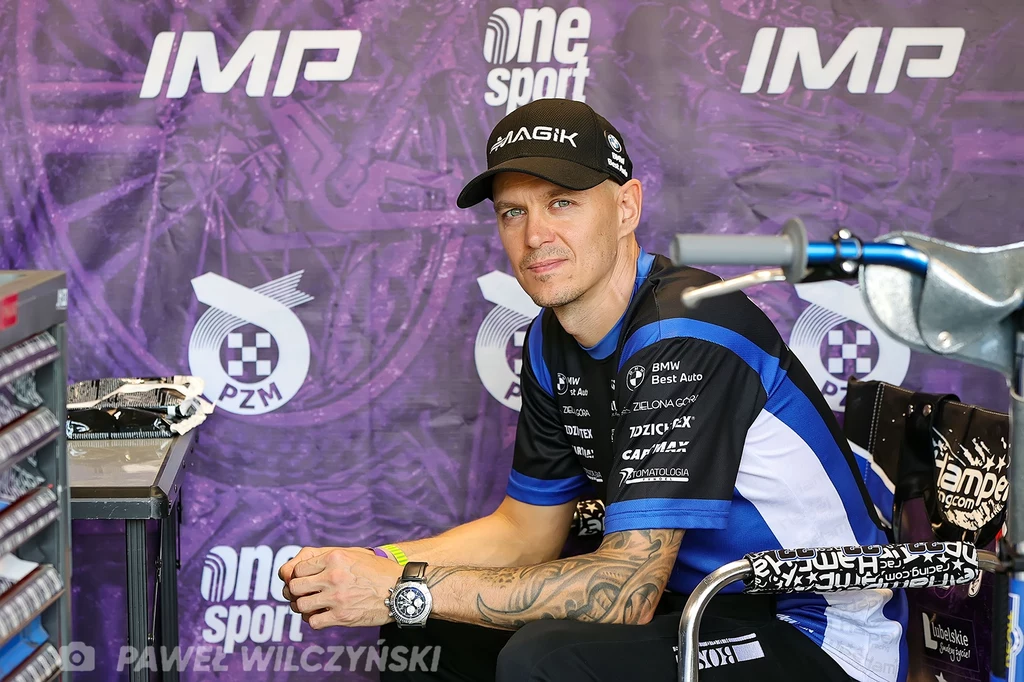 Zawodnik siedzący w boksie żużlowym, ubrany w sportową koszulkę i czapkę z daszkiem, na tle baneru z logo IMP oraz One Sport, z widocznym fragmentem motocykla po prawej stronie.