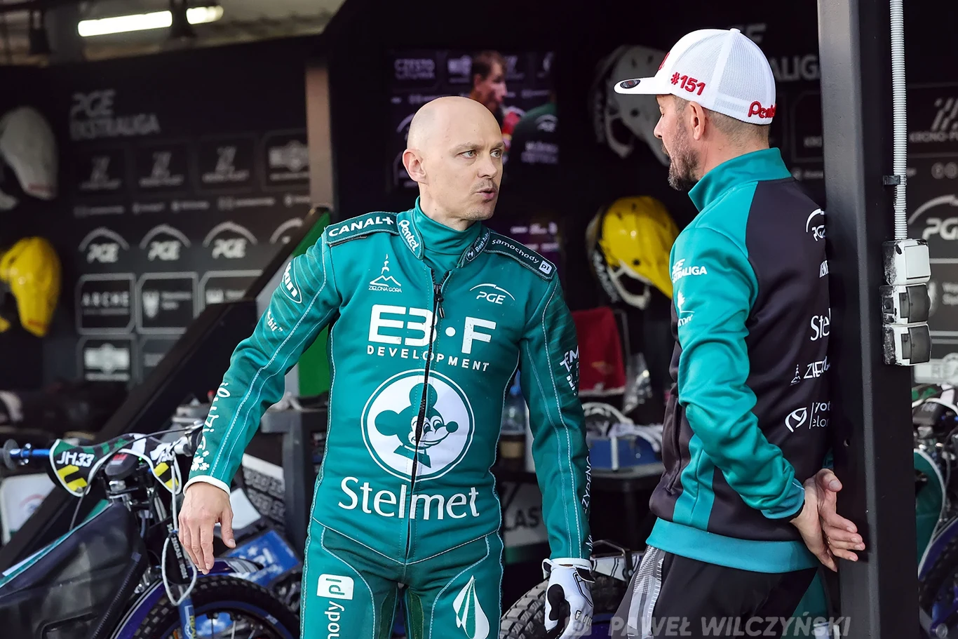 Dwóch mężczyzn w zielonych sportowych kombinezonach rozmawia w boksie żużlowym, w tle widoczne motory, części zamienne oraz tablice z logotypami sponsorów.