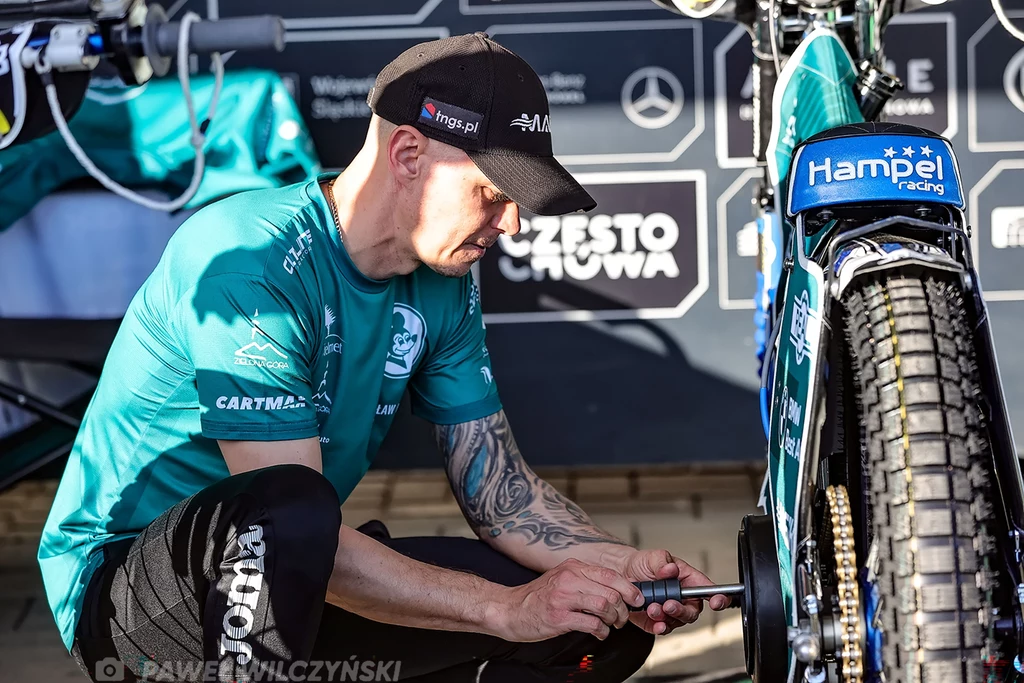 Mechanik ubrany w zieloną koszulkę serwisuje motocykl żużlowy, skupiając się na elementach tylnego koła, w tle widoczny baner sponsorski i narzędzia serwisowe.