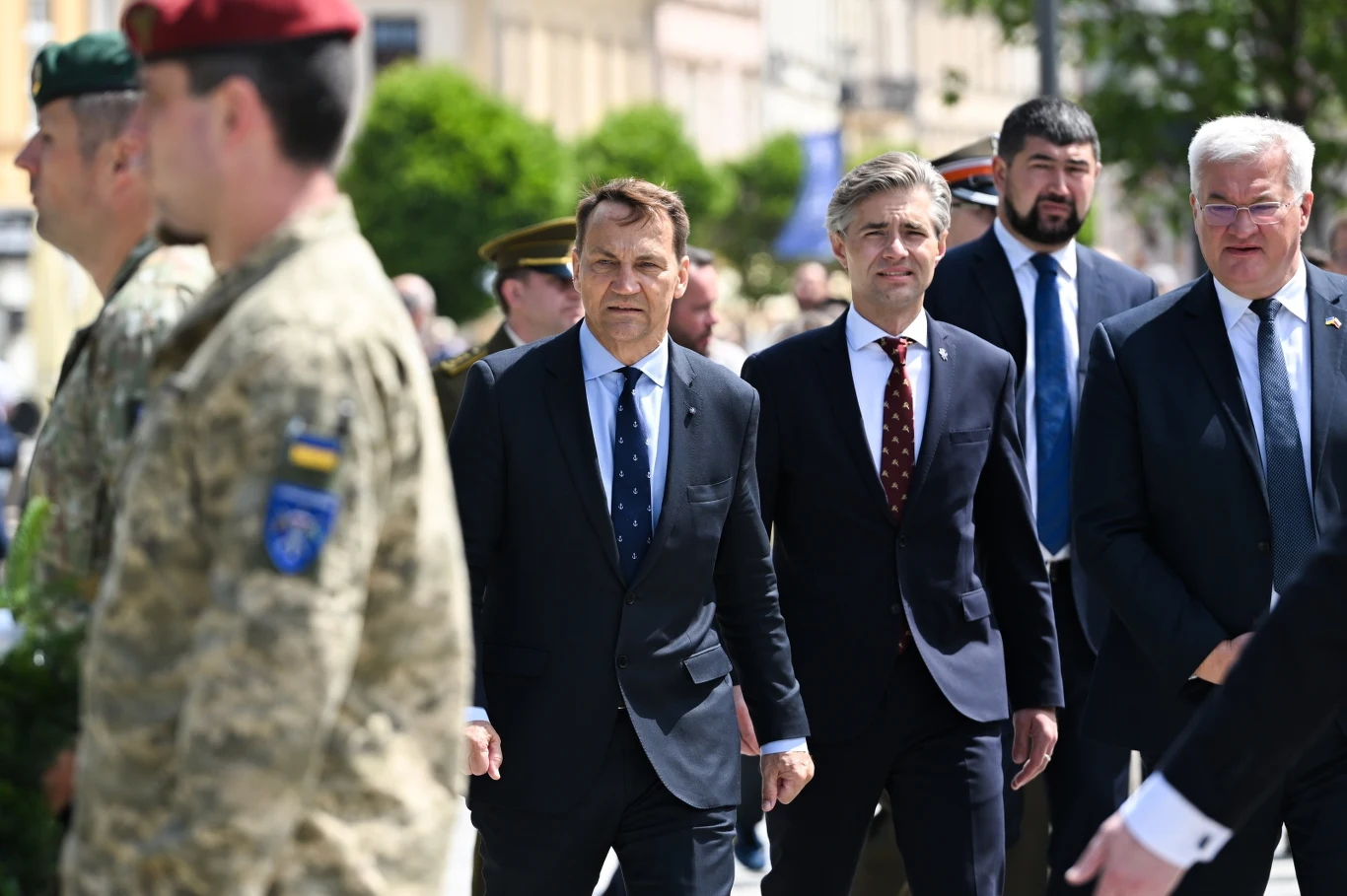 Szef MSZ Radosław Sikorski w Lublinie mówił o rosyjskim ataku na polską firmę w Winnicy Uroczysty przemarsz, w centrum trzech elegancko ubranych mężczyzn w garniturach, w otoczeniu wojskowych w mundurach, sceneria zewnętrzna z zielenią i budynkami w tle.