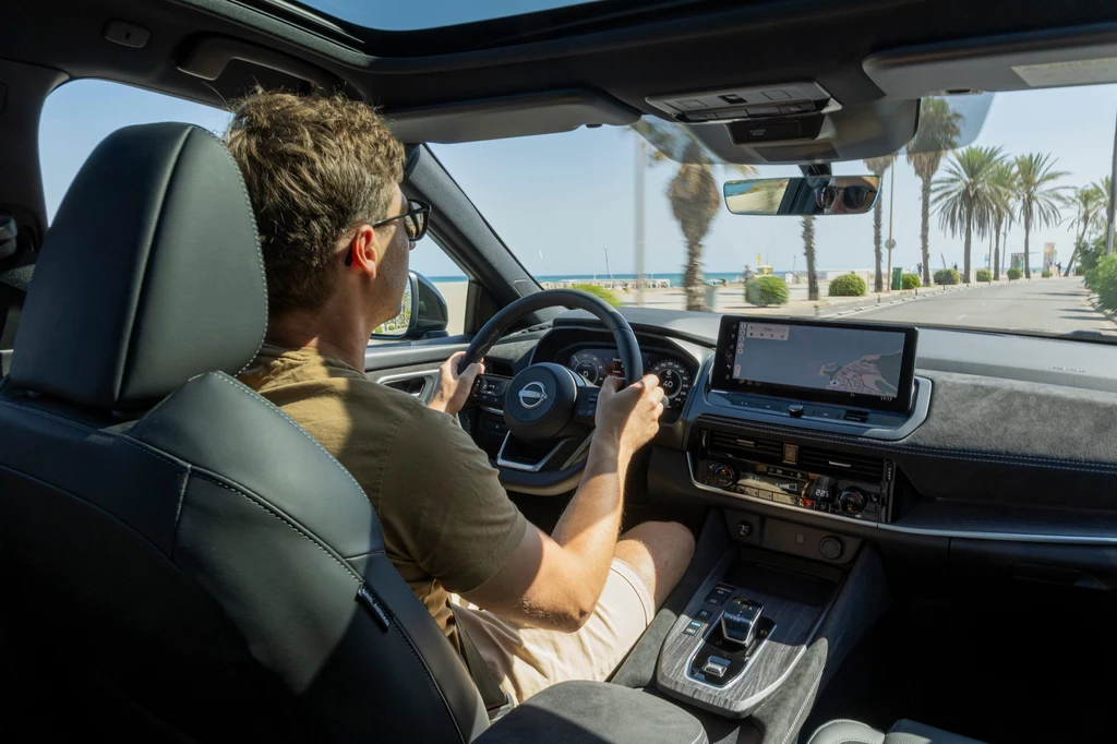 Qashqai z nowym napędem otrzymał przy okazji nowe funkcje Google w swoich multimediach – między innymi obsługę głosową Asystenta. Mężczyzna prowadzi samochód z nowoczesnym wnętrzem, szeroka droga biegnie wzdłuż palm i wybrzeża, jasne światło słoneczne wpada przez panoramiczny dach.