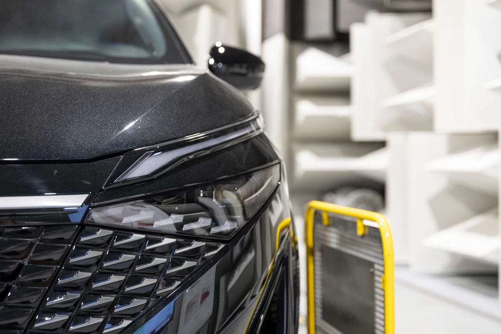 Nissan Qashqai z nowym napędem e-Power III generacji - poznasz go między innymi po czarnych obudowach lusterek. Zbliżenie na przedni reflektor oraz część maski nowoczesnego czarnego samochodu osobowego, w tle elementy labolatorium lub komory testowej z białymi panelami akustycznymi oraz żółtym elementem ochronnym.