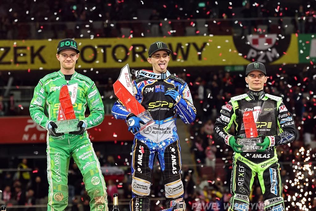 Jack Holder triumfator Grand Prix Warszawy. Obok Brady Kurtz i Patryk Dudek Trzech sportowców w kombinezonach motocyklowych stoi na podium podczas ceremonii wręczenia nagród, trzymając trofea i polskie flagi, otoczonych konfetti i światłami stadionu.