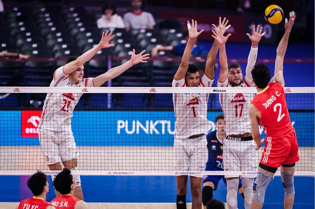 Liga Narodów siatkarzy. Kadr z meczu Chiny - Francja Zawodnik drużyny w czerwonych strojach atakuje piłkę podczas meczu siatkówki, trzech siatkarzy drużyny przeciwnej ustawia blok przy siatce, a wokół widać trybuny z widzami.