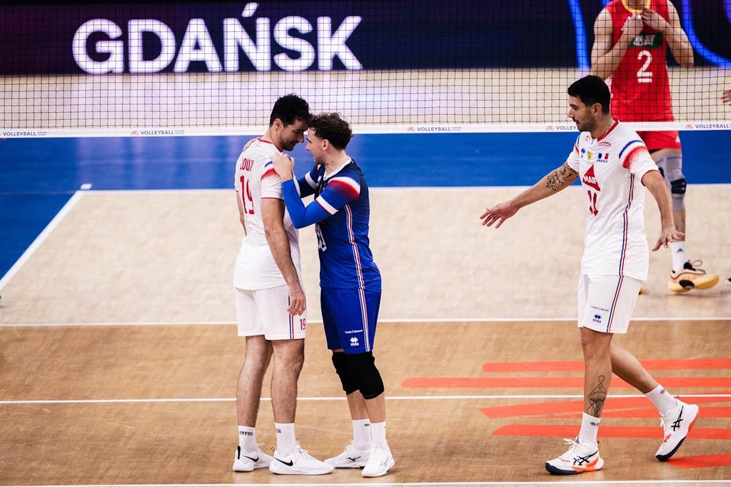 Siatkarze reprezentacji Francji Dwóch siatkarzy w białych strojach oraz jeden w niebieskim stroju na hali sportowej, jeden z zawodników dotyka ramienia drugiego w geście wsparcia, w tle widoczny napis GDAŃSK oraz fragment siatki.
