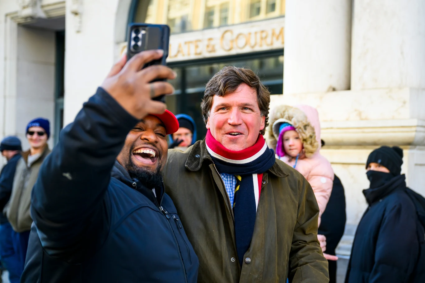 Fani robią sobie selfie z Tuckerem Carlsonem w dniu inauguracji Donalda Trumpa na prezydenta USA Dwóch mężczyzn uśmiecha się szeroko, robiąc sobie wspólne selfie na tle miejskiej ulicy, w tłumie znajdują się przechodnie ubrani w zimowe kurtki i czapki.