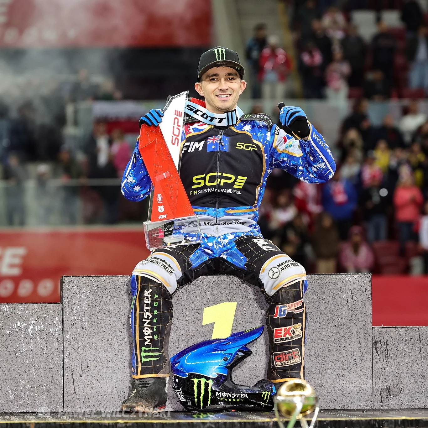Sportowiec w kombinezonie żużlowym siedzi na podium na miejscu oznaczonym cyfrą 1, trzymając trofeum w jednej ręce i mając obok siebie kask, w tle widoczni są kibice na trybunach.