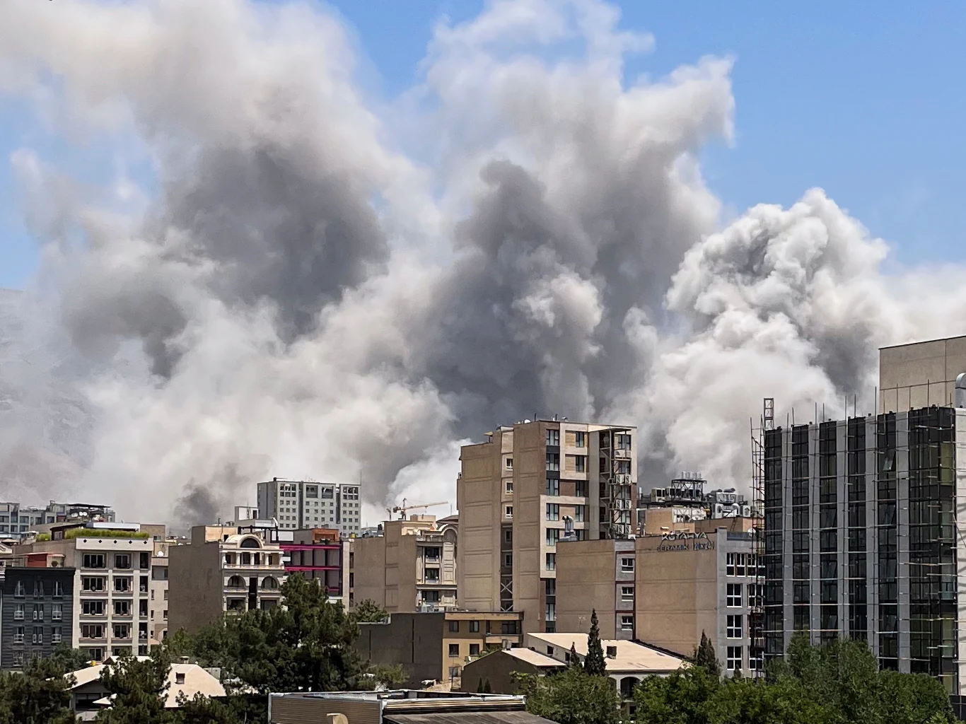 Kłęby dymu nad północną częścią Teheranu po izraelskich atakach Gęste kłęby dymu unoszące się nad zabudowaniami miejskimi, otoczone budynkami mieszkalnymi i biurowymi w jasny, słoneczny dzień. Dym całkowicie dominuje na tle nieba, tworząc atmosferę niepokoju i zagrożenia.