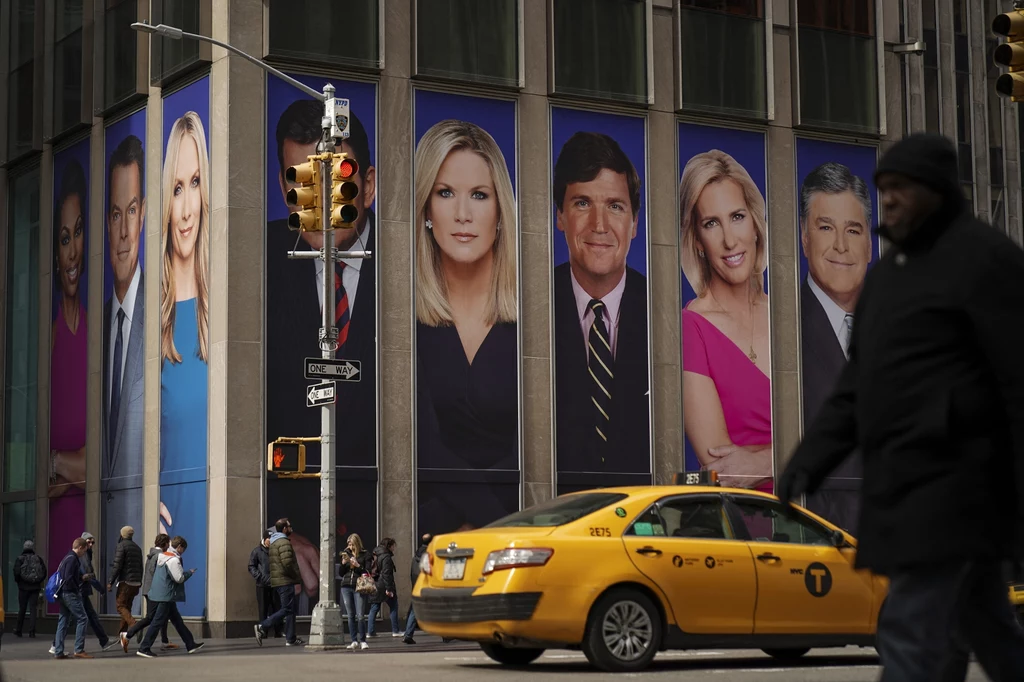 Baner z twarzami dziennikarzy Fox News, w tym Tuckera Carlsona, na budynku News Corporation w Nowym Jorku Wielkoformatowe portrety znanych prezenterów telewizyjnych na fasadzie nowoczesnego budynku w centrum miasta, poniżej żółta taksówka przejeżdża przez skrzyżowanie, na chodniku kilku pieszych.