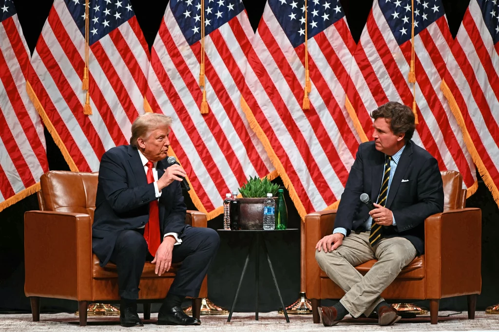 Wywiad Tuckera Carlsona z Donaldem Trumpem pod koniec października 2024 r. Dwóch mężczyzn siedzi na fotelach podczas rozmowy na scenie, w tle ustawiony rząd amerykańskich flag, między nimi stolik z butelką wody i rośliną, obaj trzymają mikrofony.