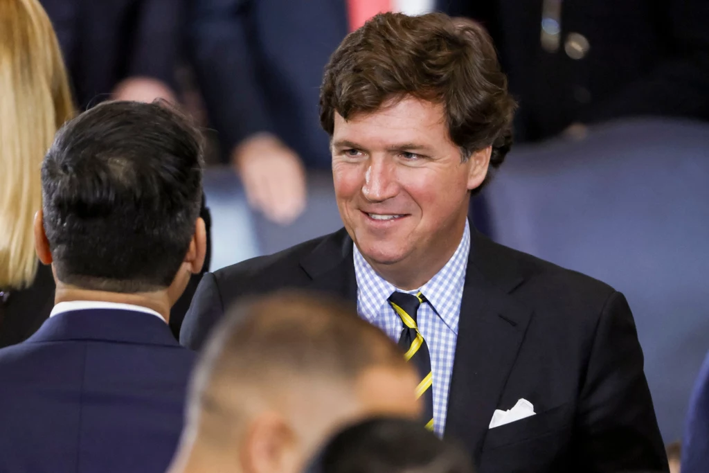 Tucker Carlson podczas ceremonii zaprzysiężenia Donalda Trumpa na prezydenta USA Mężczyzna w garniturze z żółto-czarnym krawatem uśmiecha się i rozmawia z innymi osobami na tle grupy ludzi.