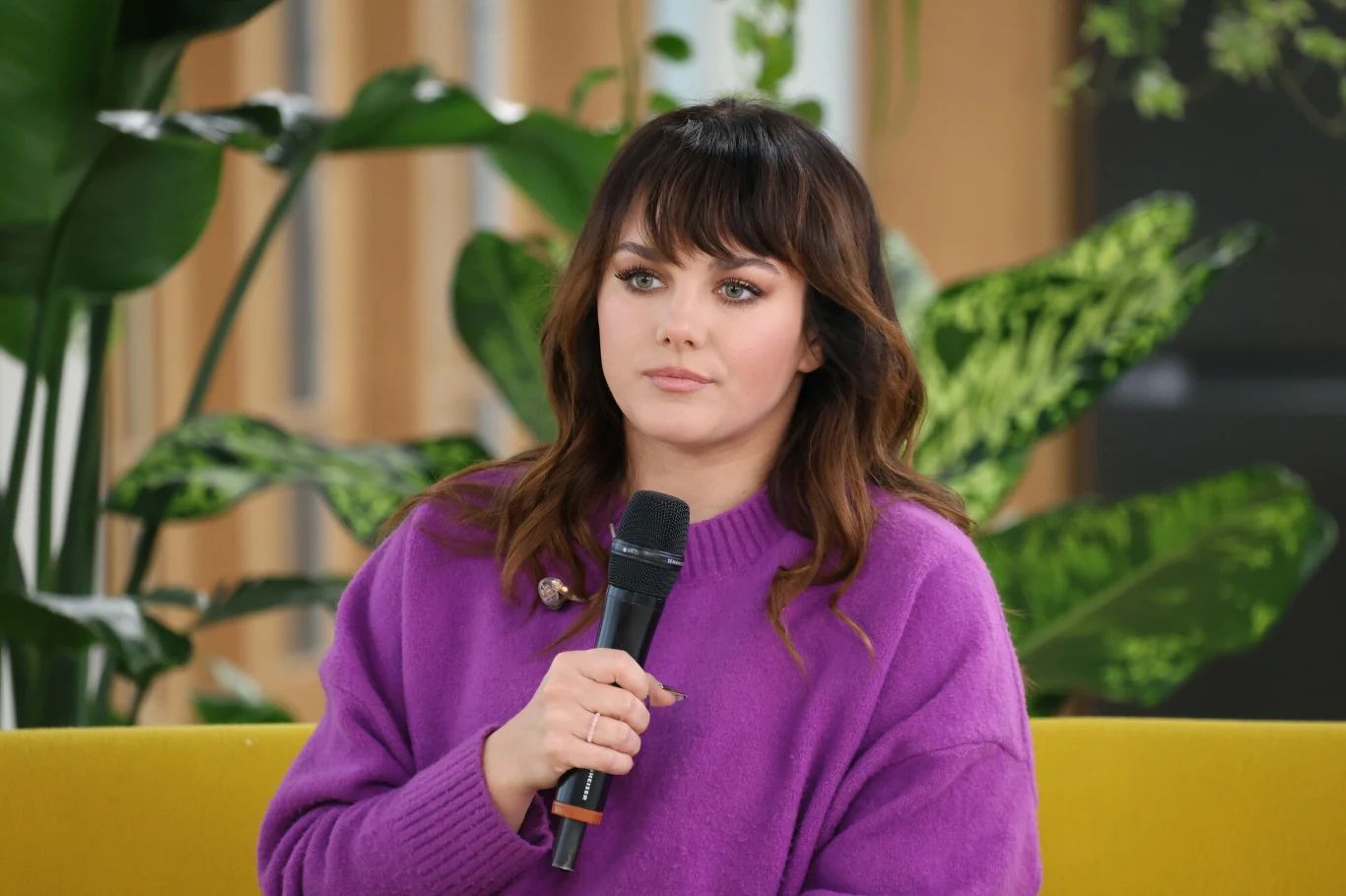 Ewa Farna wyruszy w wyjątkową trasę koncertową Młoda kobieta z ciemnymi włosami i grzywką, ubrana w fioletowy sweter, siedzi na żółtej sofie, trzymając mikrofon. W tle widoczne zielone liście roślin doniczkowych.