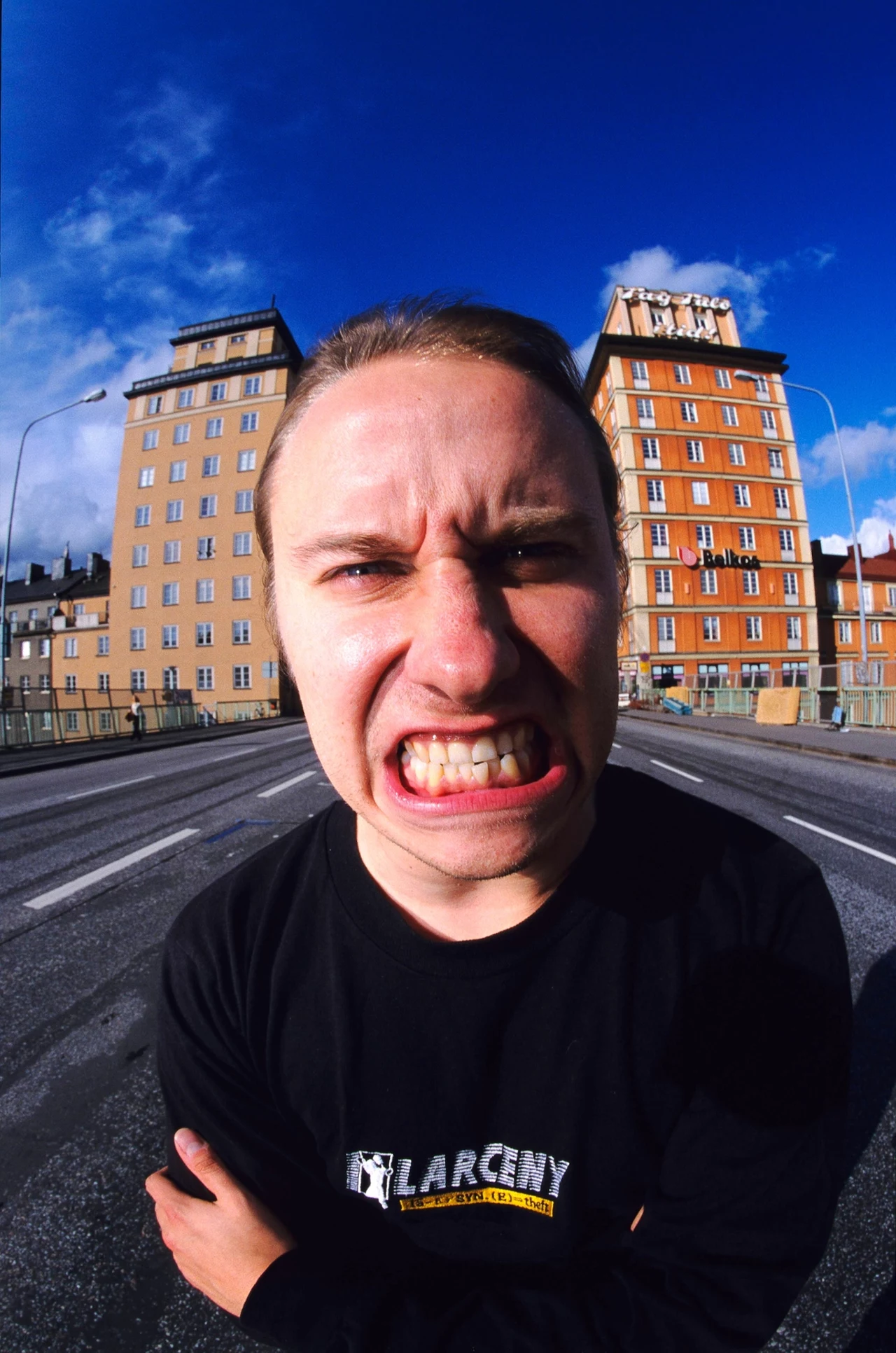 Lars-Göran ”L-G” Petrov (Entombed, Entombed A.D., Firespawn) zmarł 7 marca 2021 r. Młody mężczyzna w czarnej koszulce stoi na środku jezdni, z wykrzywioną miną i szeroko otwartymi ustami, na tle dwóch wysokich budynków oraz nieba z białymi chmurami.