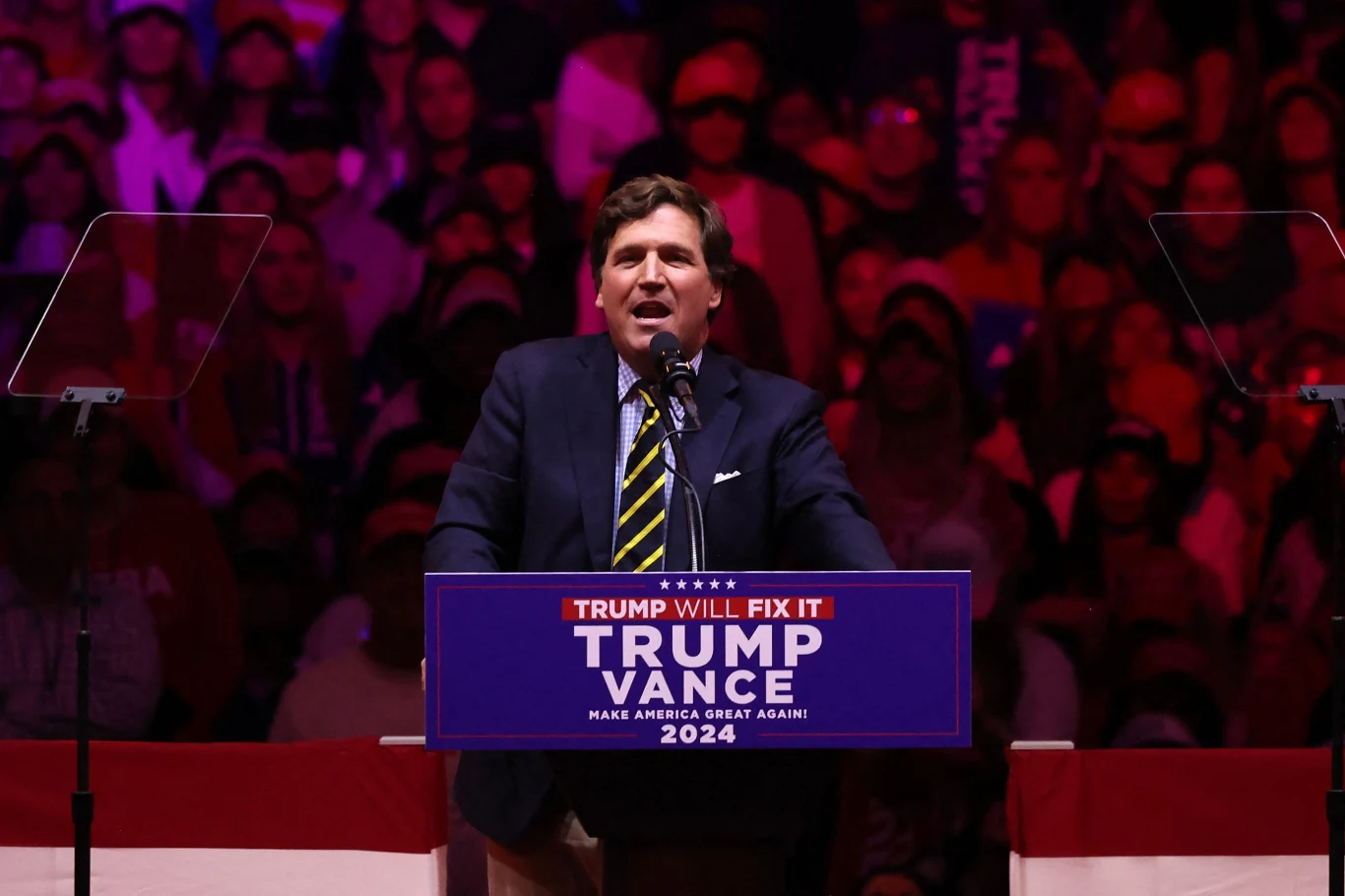 Tucker Carlson przemawia podczas wiecu Donalda Trumpa w Nowym Jorku Mężczyzna przemawia do mikrofonu na tle licznego tłumu na wiecu politycznym, przed pulpitem z napisem Trump Vance 2024 oraz hasłem Trump Will Fix It, wnętrze oświetlone mocnym czerwonym światłem, w tle widoczni ludzie.
