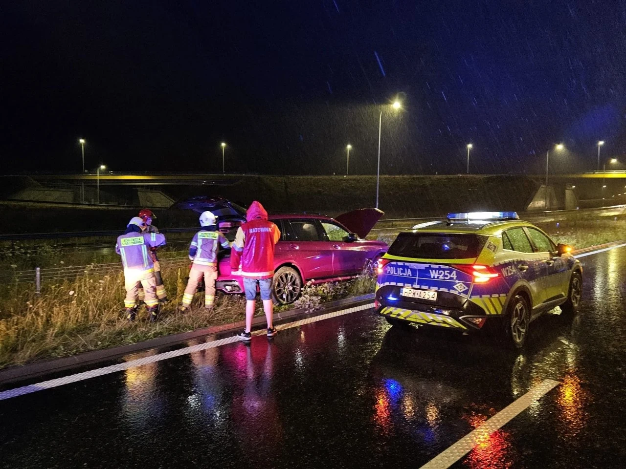 Policjanci oraz strażacy pracują przy czerwonym samochodzie stojącym na poboczu mokrej, oświetlonej drogi nocą, obok policyjny radiowóz z włączonymi światłami.