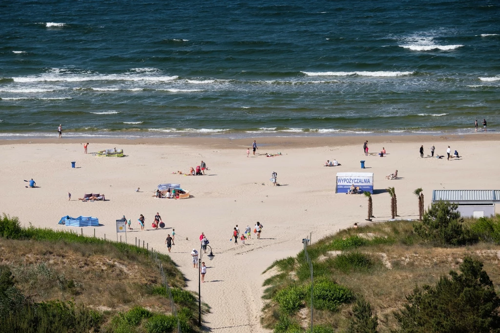 Plaża w Świnoujściu skradła serca turystów Piaszczysta plaża nad morzem z pojedynczymi grupami ludzi korzystającymi z uroków letniego wypoczynku, w tle spokojne fale oraz błękitne niebo, wejście prowadzi do szerokiej plaży o luźno rozłożonych leżakach i ręcznikach.