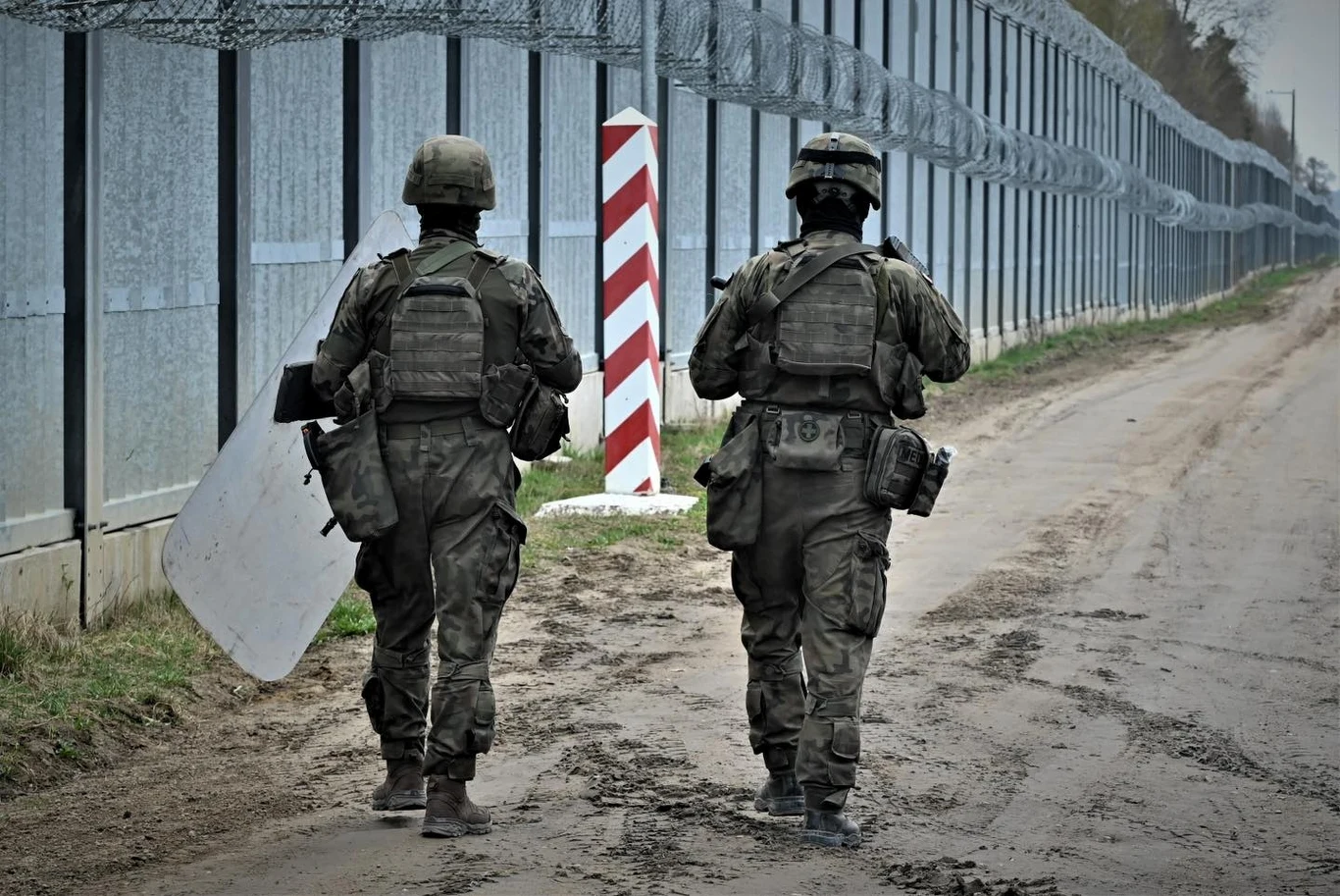 Patrol żołnierzy na polsko-białoruskiej żołnierzy Dwóch żołnierzy w pełnym umundurowaniu patroluje piaszczystą drogę biegnącą wzdłuż ogrodzenia z drutem kolczastym, obok charakterystycznego słupa granicznego w biało-czerwone pasy.