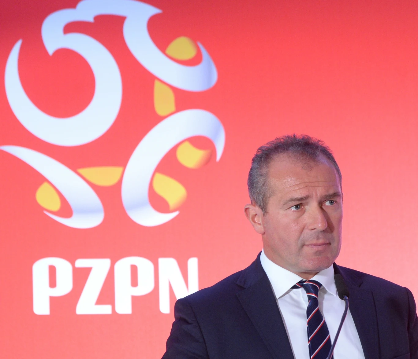 Mężczyzna w garniturze i krawacie, stojący przed czerwonym tłem z dużym logo Polskiego Związku Piłki Nożnej oraz napisem PZPN.
