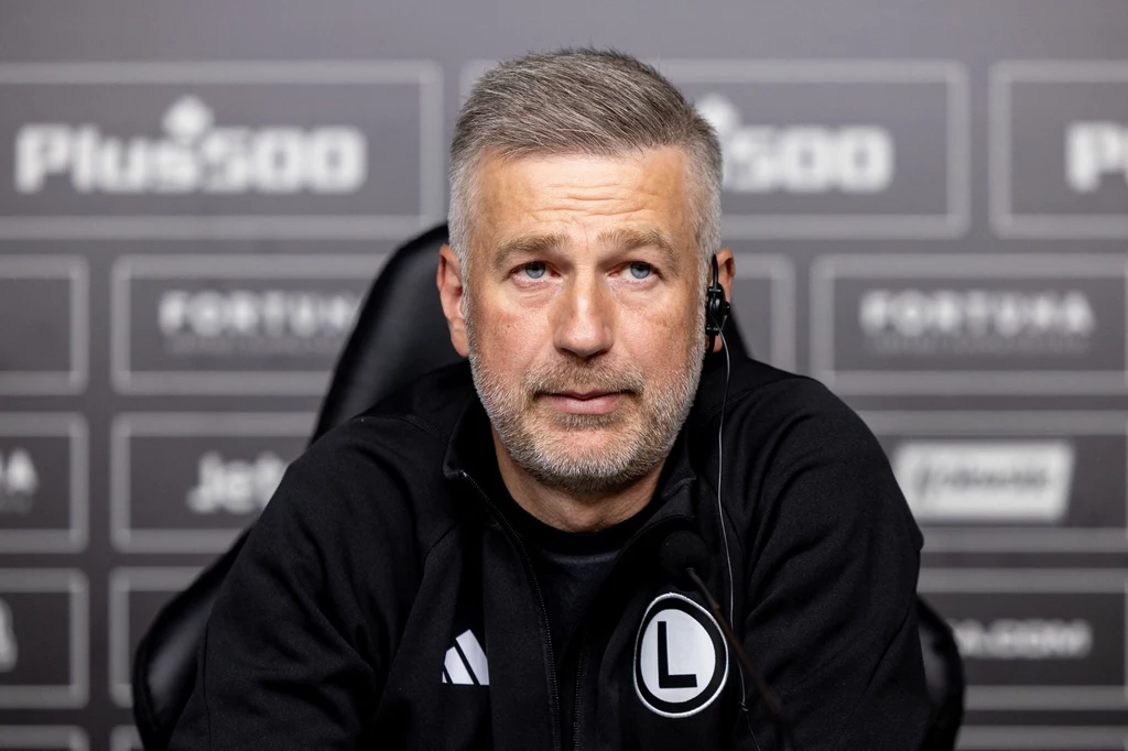 Edward Iordanescu Mężczyzna w czarnej bluzie sportowej z logo klubu Legia Warszawa, siedzi na tle ściany z logotypami sponsorów podczas konferencji prasowej. Na jego uchu widoczna jest słuchawka.