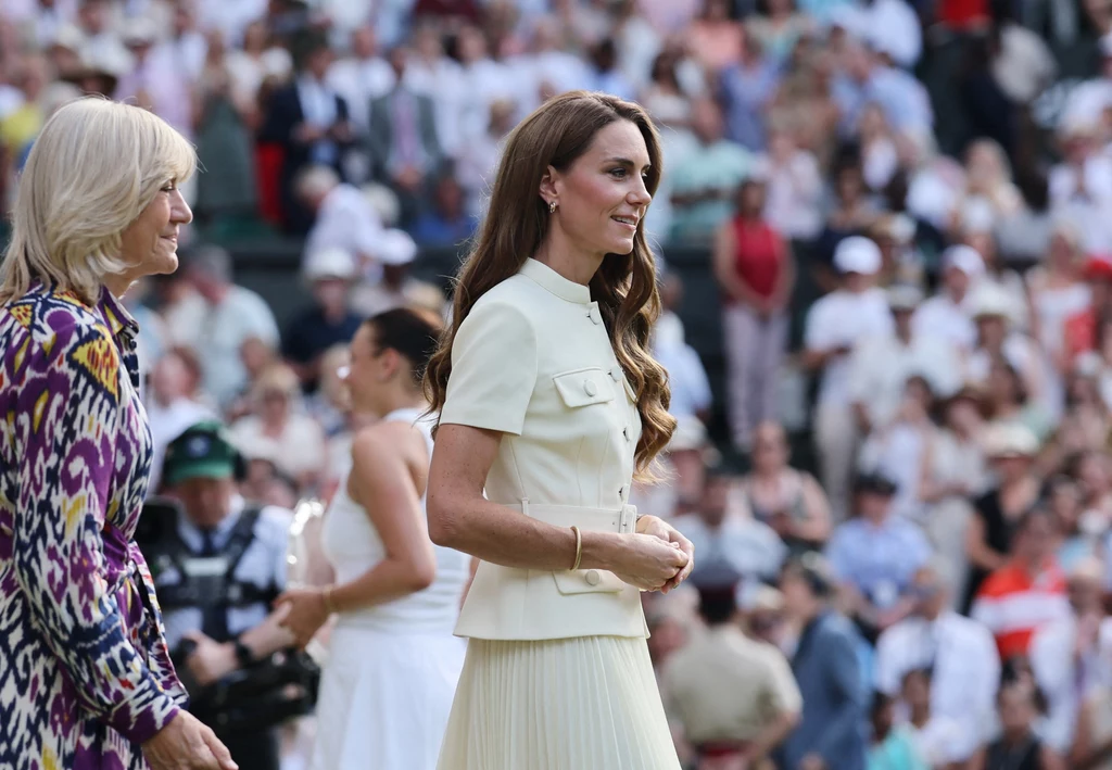 Księżna Kate, Wimbledon 2025 Elegancko ubrana kobieta w jasnej stylizacji stoi na tle tłumu ludzi, u jej boku kobieta w kolorowej marynarce, w tle widać zebranych widzów i zawodniczkę na korcie sportowym.