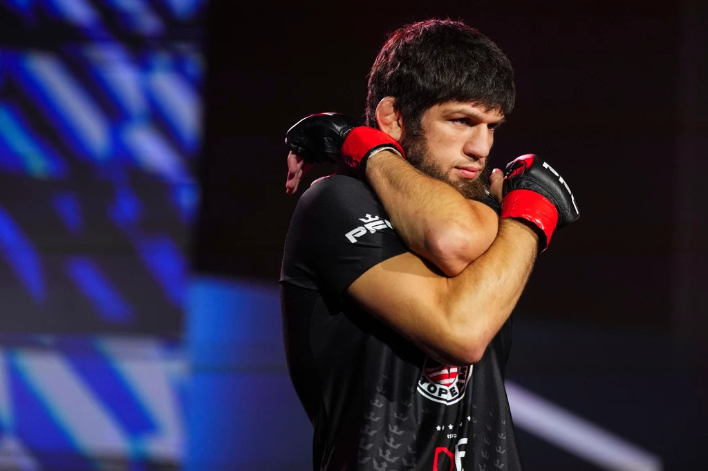 Timur Khizriev Zawodnik MMA w czarnej koszulce i rękawicach sportowych, skoncentrowany i przygotowujący się do walki, w tle rozmyte światła areny.