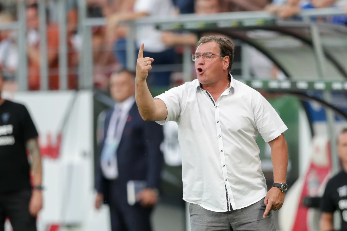 Trener w okularach i białej koszuli energicznie gestykuluje na tle stadionu, wskazując palcem wskazującym do góry, za nim rozmyte sylwetki innych osób i fragmenty trybun.