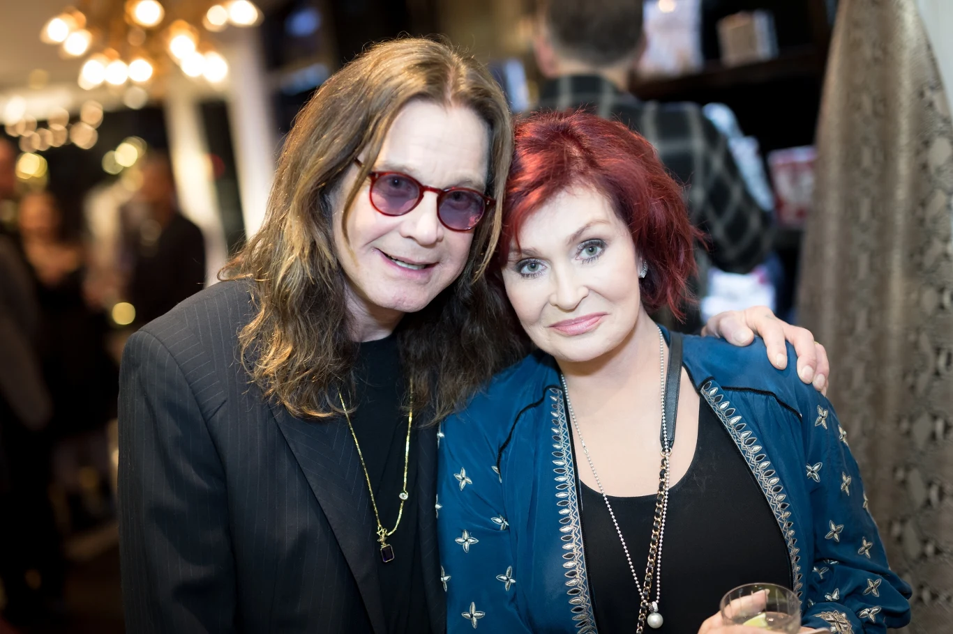 Ozzy Osbourne ma wierną rzeszę fanów, rozsianą po całym świecie Dwie uśmiechnięte osoby pozują razem w eleganckich ubraniach, jedna z długimi brązowymi włosami i okularami przeciwsłonecznymi, druga z krótkimi, czerwonymi włosami i niebieską narzutką w pomieszczeniu pełnym innych ludzi.