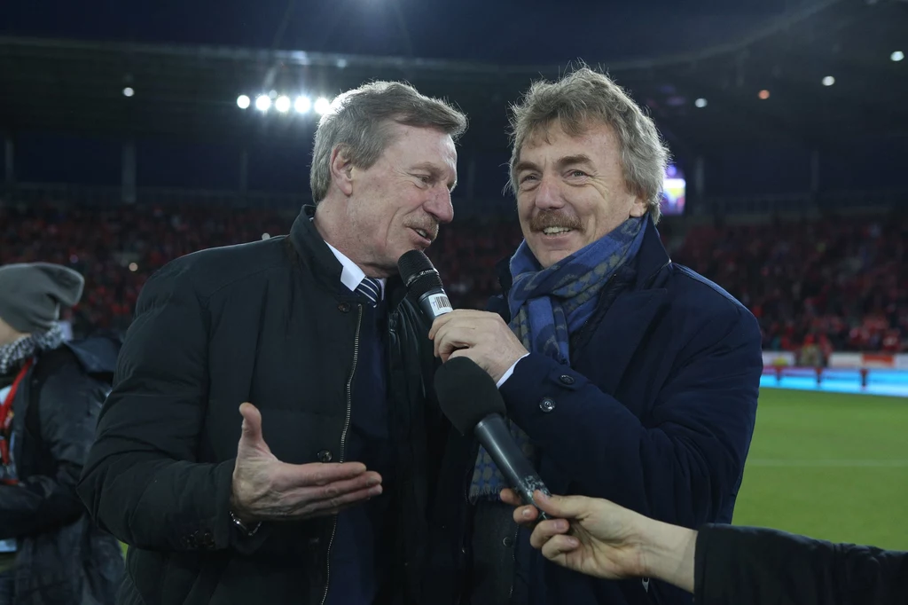 Czasy współczesne. Józef Młynarczyk i Zbigniew Boniek, dwaj przyjaciele z łódzkiego Widzewa Dwóch mężczyzn w eleganckich płaszczach i szalach rozmawia ze sobą na tle stadionu, jeden z nich trzyma mikrofon, obaj uśmiechnięci, wokół nich światła i publiczność.