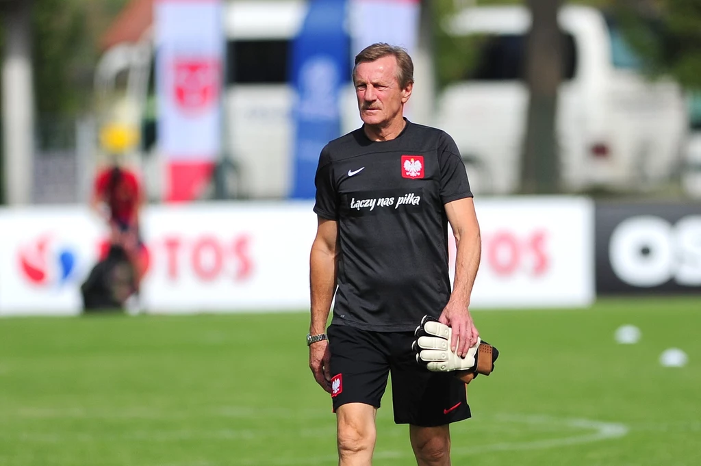 Józef Młynarczyk Mężczyzna w sportowym stroju z herbem Polski i napisem 'Łączy nas piłka' na koszulce, trzymający rękawice bramkarskie, spaceruje po trawiastym boisku piłkarskim podczas treningu.