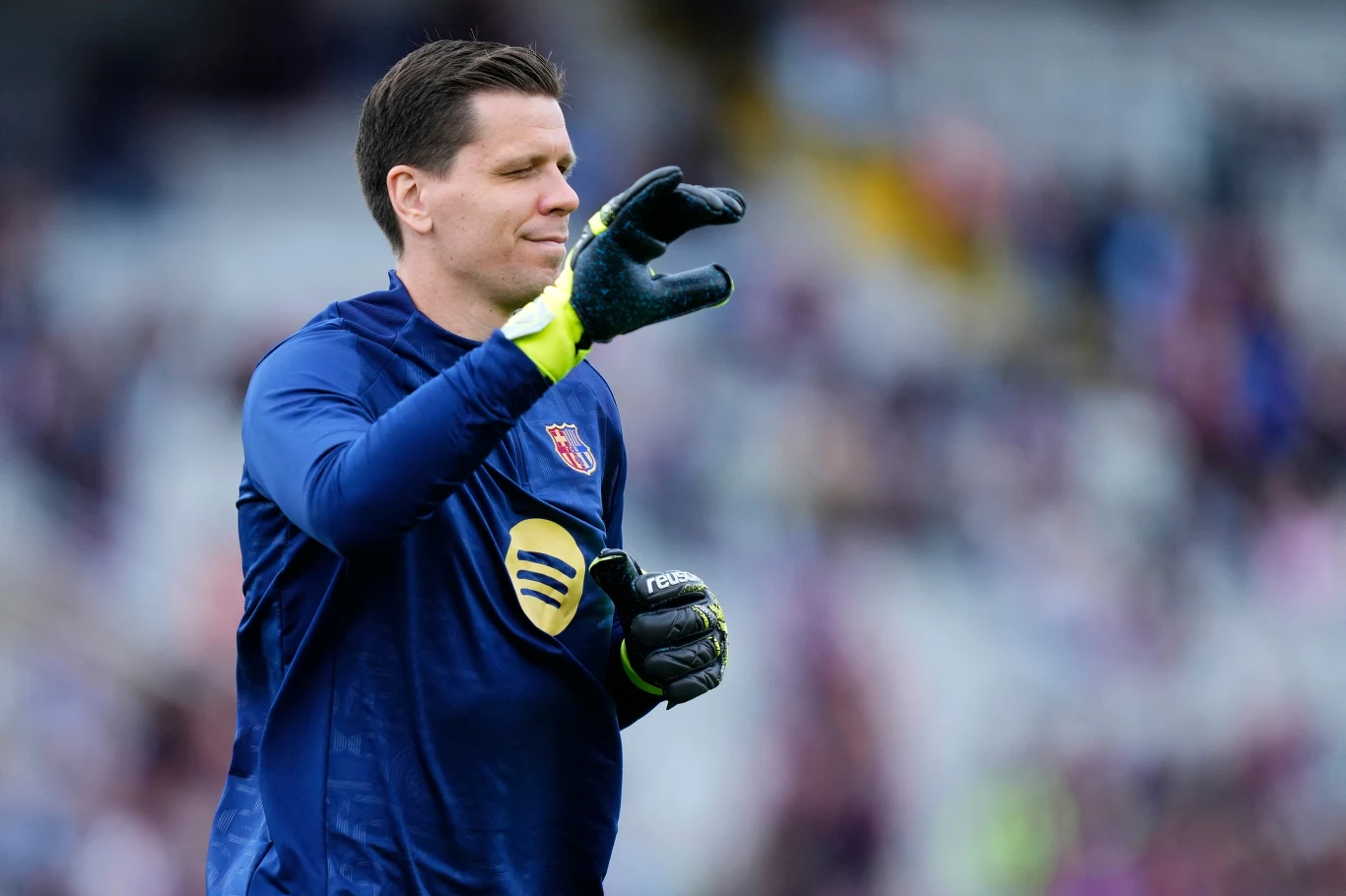 Bramkarz w niebieskim stroju z rękawicami, wykonujący gest dłonią na tle rozmytej publiczności stadionu, skupiony przygotowuje się do gry.