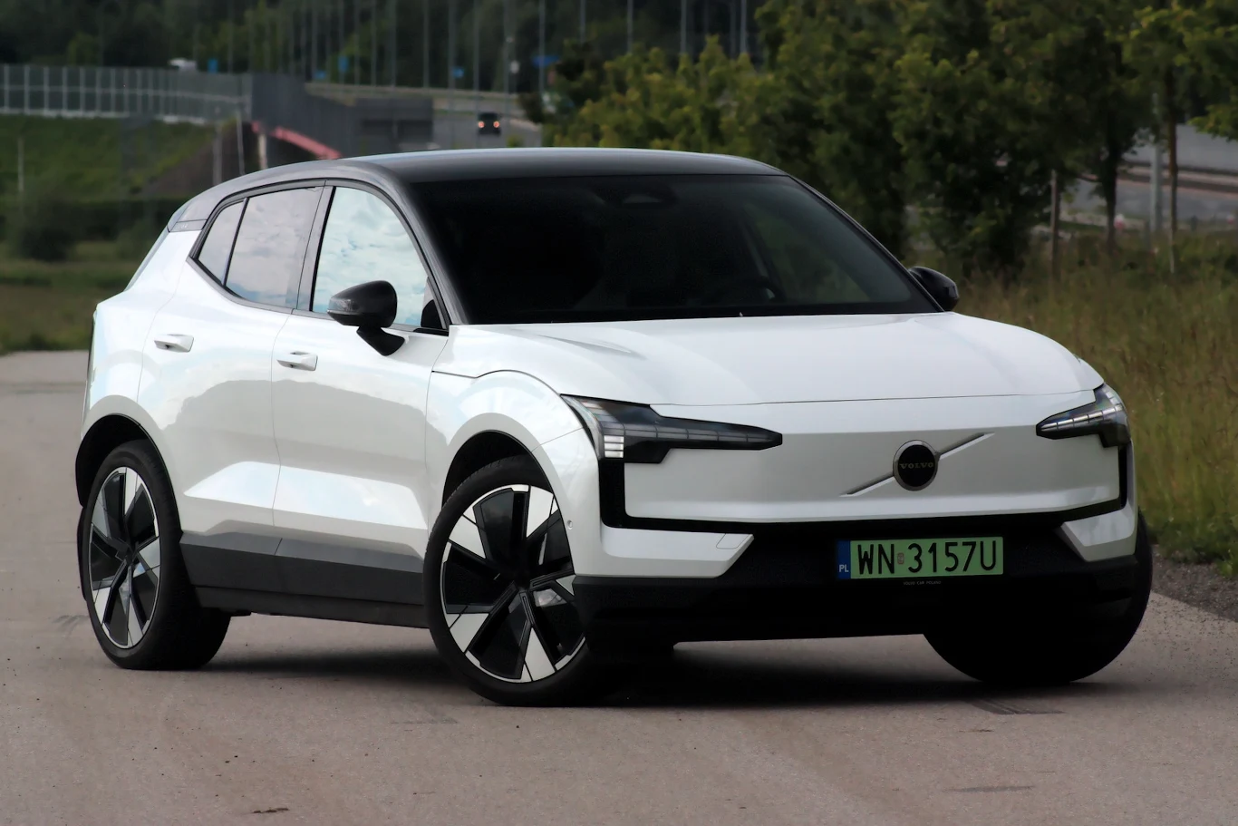 Volvo EX30 to połączenie szwedzkiego stylu i chińskiej technologii Nowoczesny biały SUV z czarnym dachem i dużymi felgami stoi na drodze, w tle zieleń i rozmyte elementy infrastruktury drogowej.