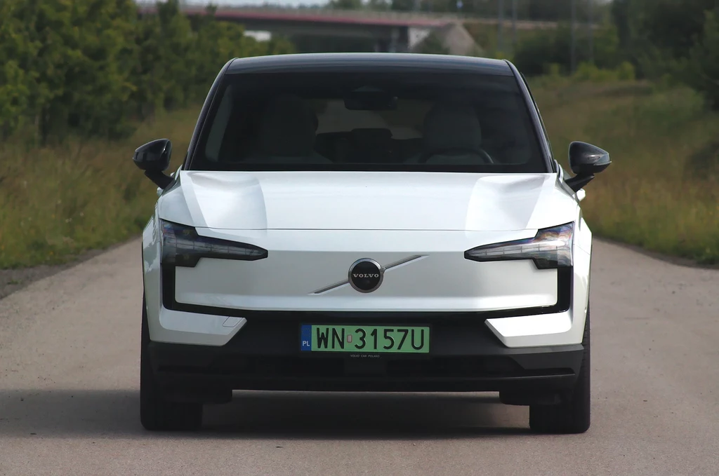 Z przodu w oczy rzucają się zabudowana pas przedni i charakterystyczne światła Nowoczesny, biały samochód elektryczny marki Volvo, widziany od frontu, stoi na asfaltowej drodze otoczonej zielenią, z wyraźnie widoczną tablicą rejestracyjną.