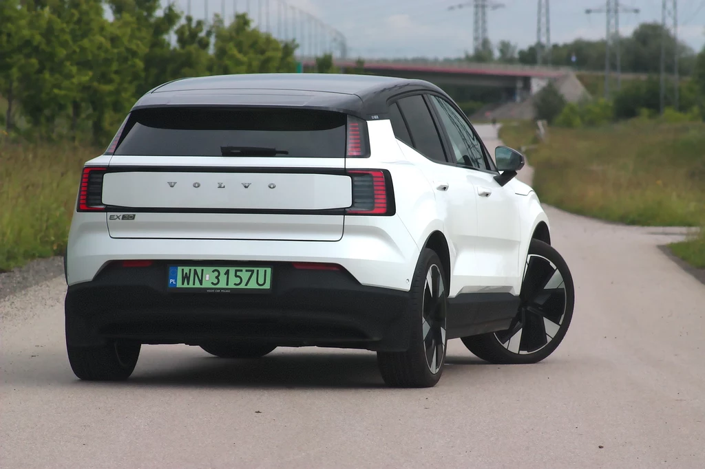 Volvo EX30 również z tyłu nie jest ekstrawaganckie Biały samochód elektryczny Volvo EX30 z czarnym dachem jedzie pustą, asfaltową drogą w otoczeniu zieleni, z widocznym mostem w tle.