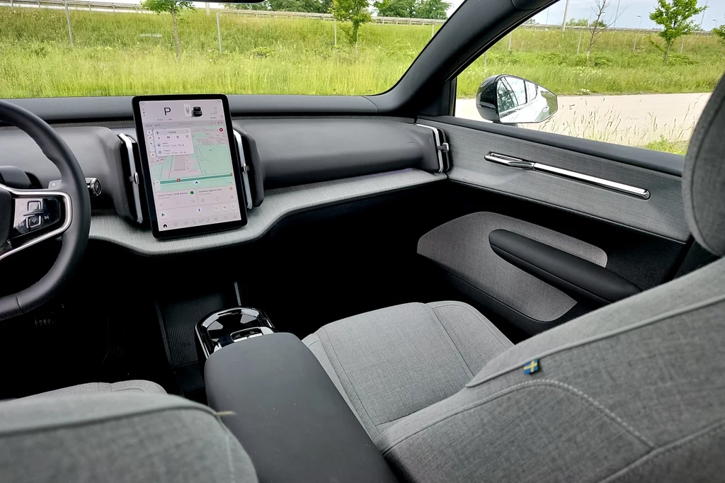 Kabina Volvo EX30 pełna jest ekologicznych materiałów z odzysku Nowoczesne, minimalistyczne wnętrze samochodu elektrycznego z dużym ekranem dotykowym na centralnej konsoli, jasnoszare fotele z tkaniny, widoczny fragment kierownicy oraz podłokietnik, widok na zieleń za oknem auta.
