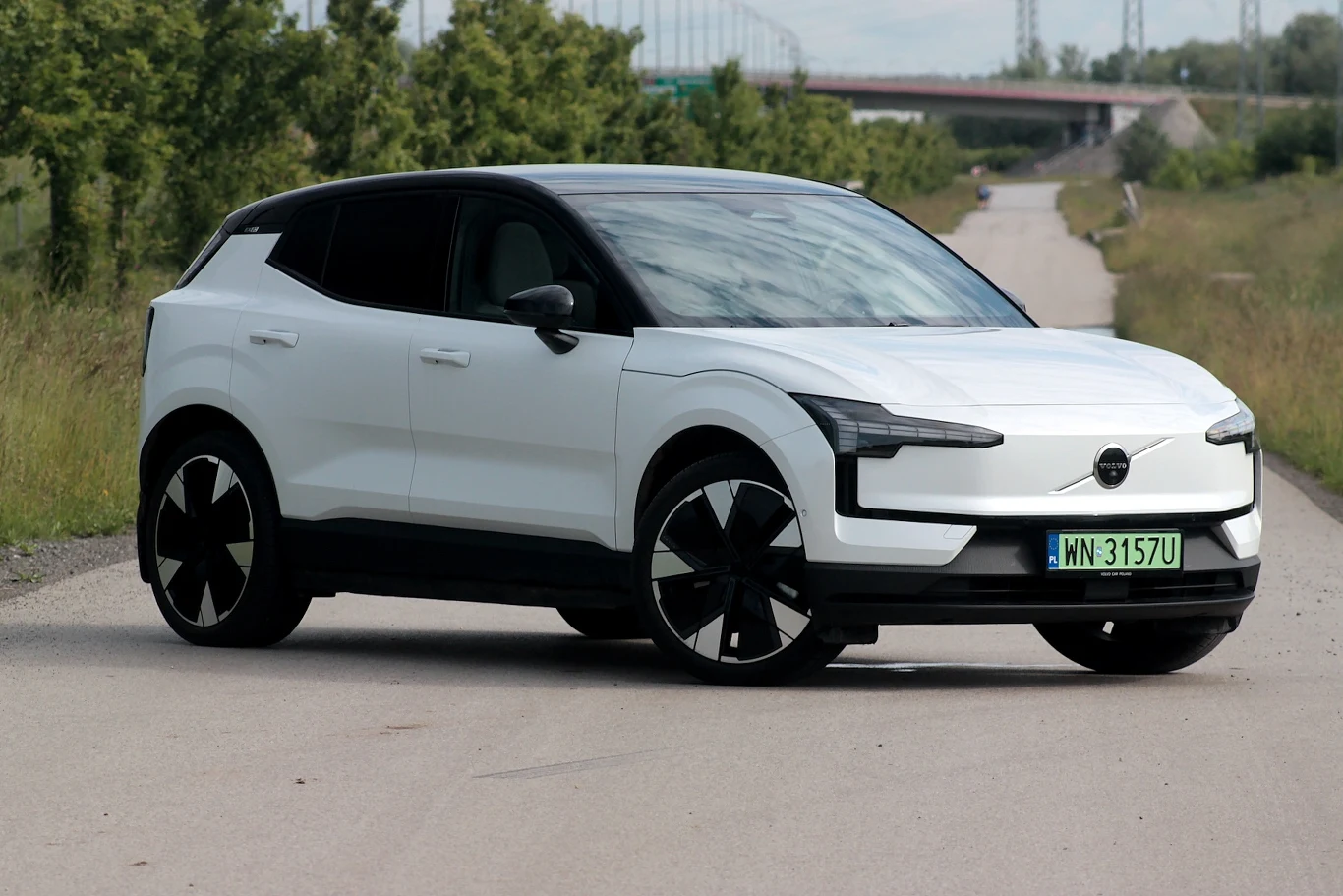Volvo EX30 Biały nowoczesny samochód elektryczny typu SUV stoi na pustej asfaltowej drodze z zielenią po bokach i mostem w tle.