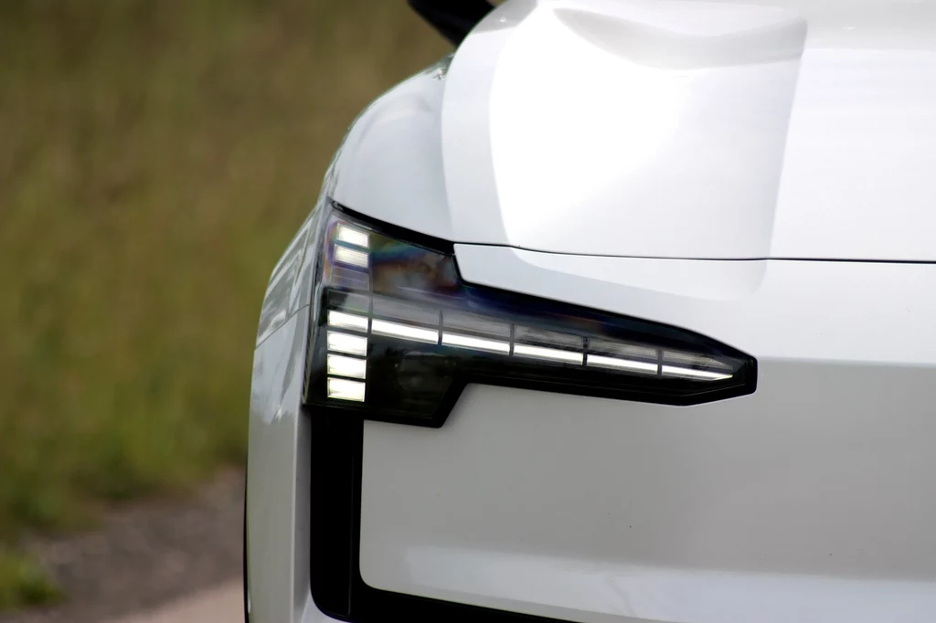 Charakterystyczne dla wszystkich modeli Volvo są światłem z motywem młota Thora Przedni reflektor nowoczesnego białego samochodu z wyraźnie widocznymi światłami LED i aerodynamiczną linią maski, fragment karoserii oraz tło rozmazane, zielone.