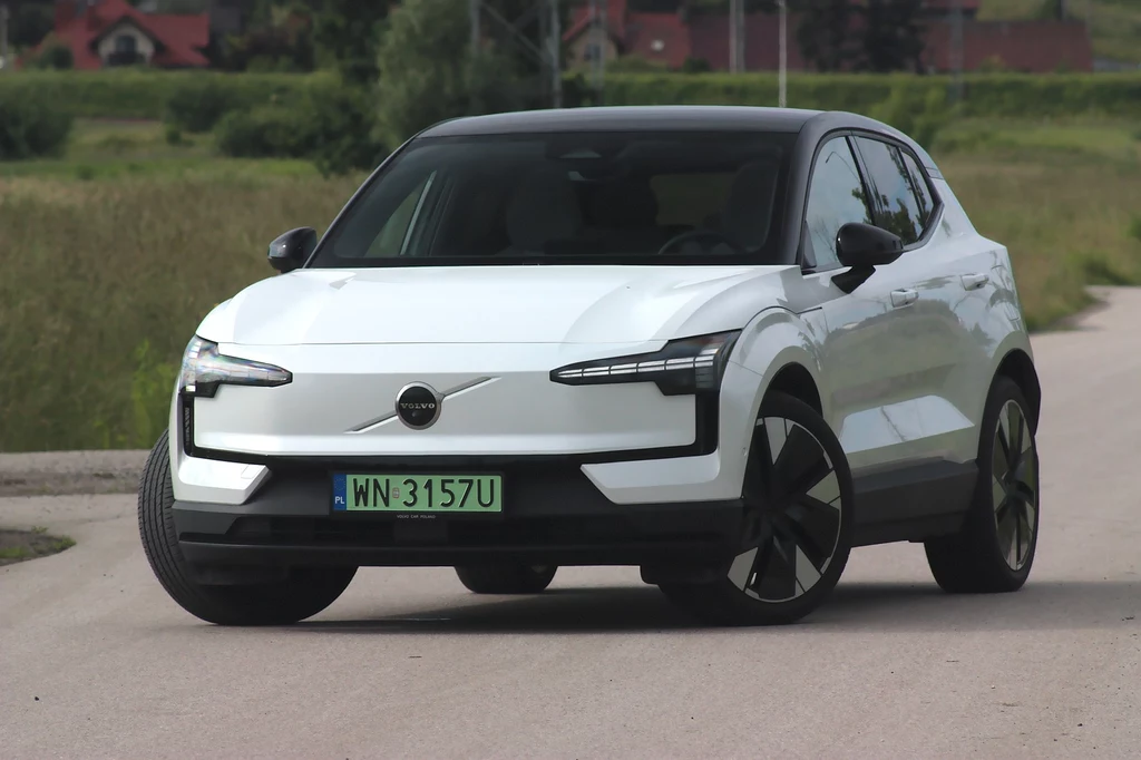 Volvo EX30 to samochód głównie dla dwóch osób Nowoczesny biały samochód elektryczny Volvo stoi na wiejskiej drodze, otoczony zielonymi polami. Samochód przyciąga uwagę charakterystycznym wzornictwem świateł LED i czarnym dachem.