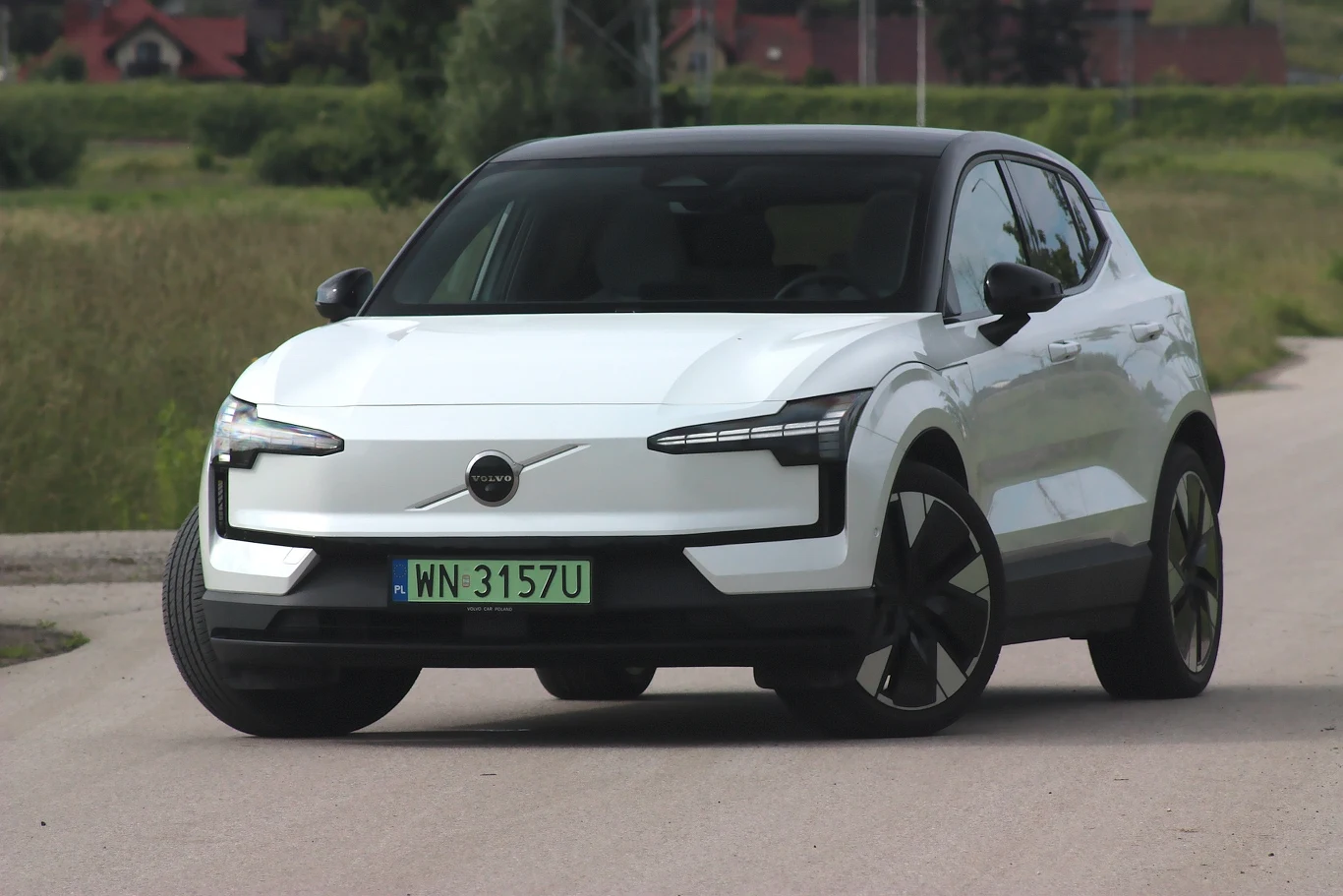 Volvo EX30 to samochód głównie dla dwóch osób Nowoczesny biały samochód elektryczny Volvo stoi na wiejskiej drodze, otoczony zielonymi polami. Samochód przyciąga uwagę charakterystycznym wzornictwem świateł LED i czarnym dachem.