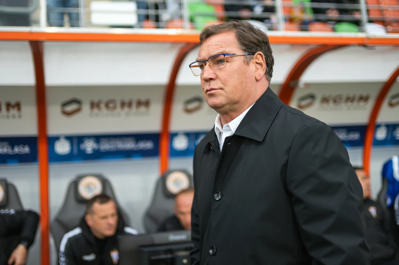 Jan Urban Mężczyzna w okularach i czarnym płaszczu, stojący na tle ławki trenerskiej podczas meczu piłkarskiego, w tle siedzą trenerzy lub członkowie sztabu szkoleniowego.