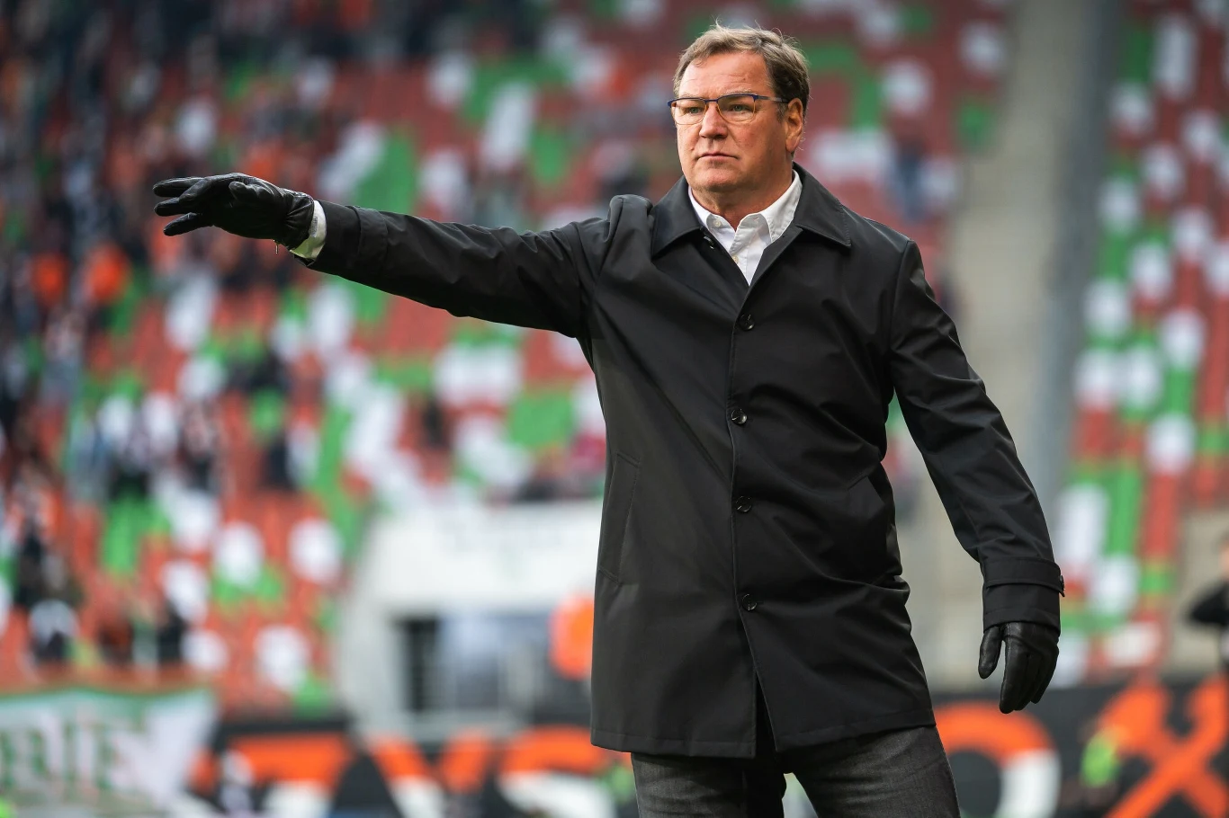 Jan Urban Mężczyzna w czarnym płaszczu i rękawiczkach stoi na stadionie, wskazując ręką na coś poza kadrem, w tle rozmyte, kolorowe trybuny z flagami kibiców.