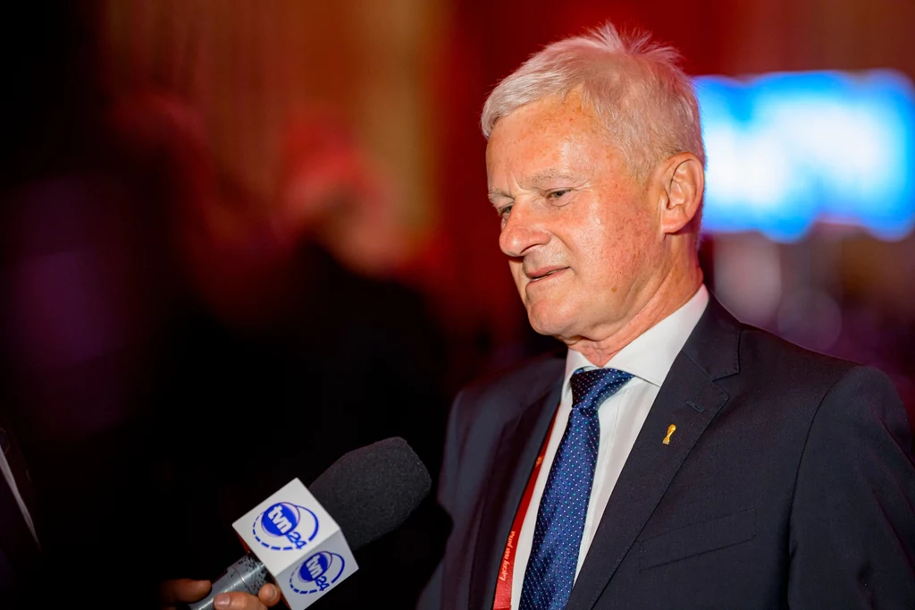 Doświadczony mężczyzna w garniturze udziela wywiadu, trzymając mikrofon z logo stacji telewizyjnej w otoczeniu ciepłego, lekko rozmytego oświetlenia.