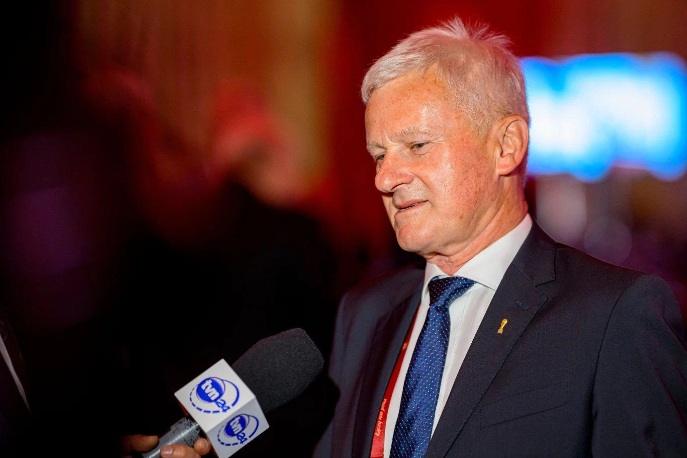 Doświadczony mężczyzna w garniturze udziela wywiadu, trzymając mikrofon z logo stacji telewizyjnej w otoczeniu ciepłego, lekko rozmytego oświetlenia.
