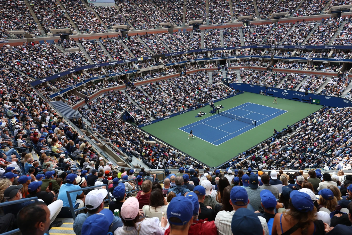 Arhur Ashe Stadium - główna arena zmagań podczas US Open Duży stadion tenisowy wypełniony kibicami, na korcie trwa mecz, a widownie otacza kort z każdej strony, tworząc imponującą i dynamiczną atmosferę sportową.