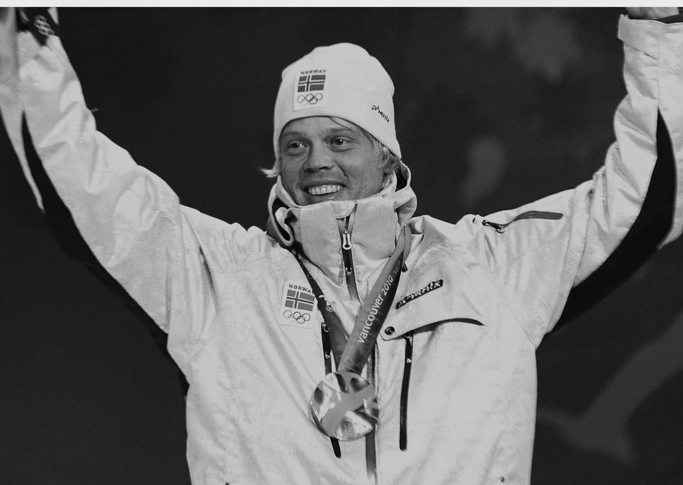 Sportowiec w zimowej kurtce i czapce z logo olimpijskim, uśmiechający się, z medalem zawieszonym na szyi, unosi ręce w geście triumfu.