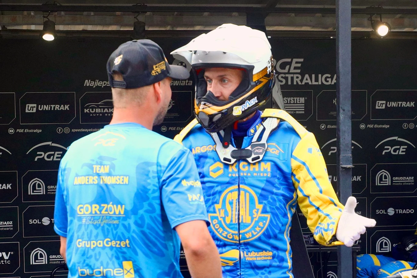 Anders Thomsen Dwóch mężczyzn ubranych w niebieskie stroje żużlowe rozmawia ze sobą w parku maszyn, jeden z nich ma założony kask motocyklowy i rękawice, wokół logotypy sponsorów i ligi żużlowej.